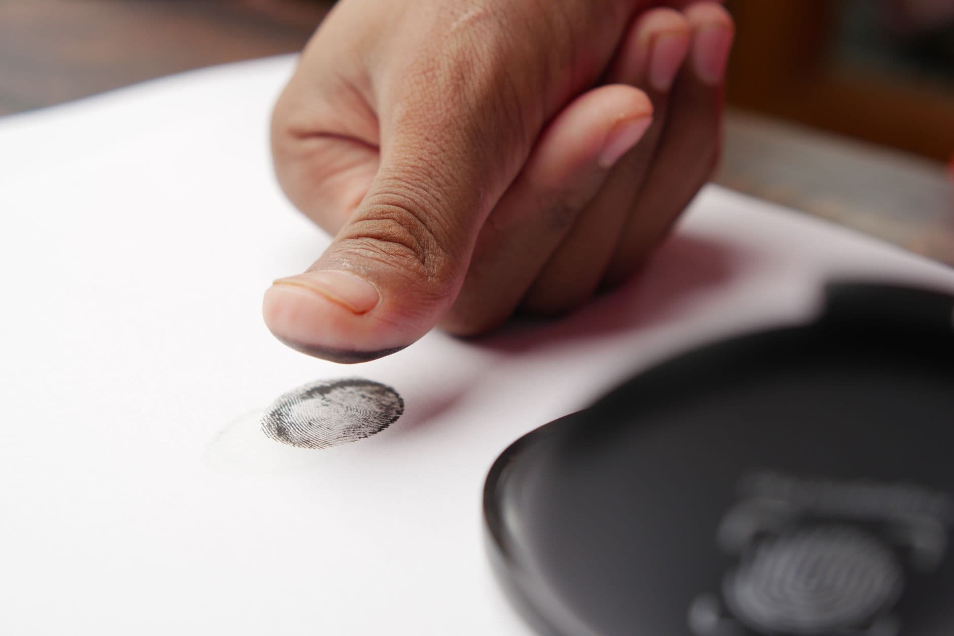 Pulgar entintado dejando una huella dactilar sobre una hoja de papel blanco, junto a un tampón de tinta.